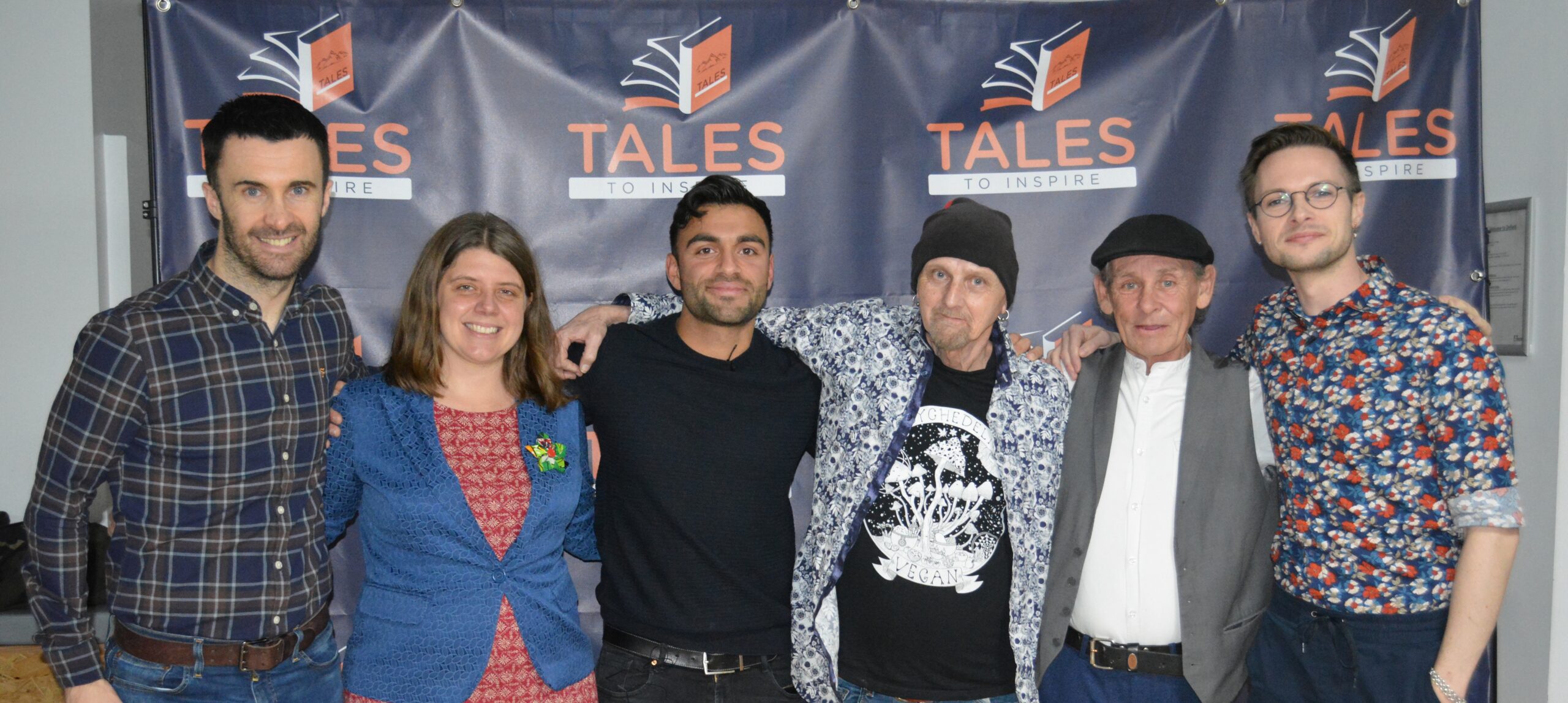 A group of 6 aduts standing in front of a Tales to Inspire banner, about to speak at a Tales to Inspire event in Manchester
